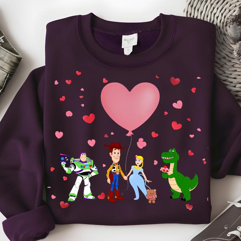 May include: A dark purple sweatshirt featuring a graphic of cartoon characters holding hands under a pink heart balloon. Red and pink hearts are scattered around the characters. The characters include a space ranger, a cowboy, a princess, and a green dinosaur.