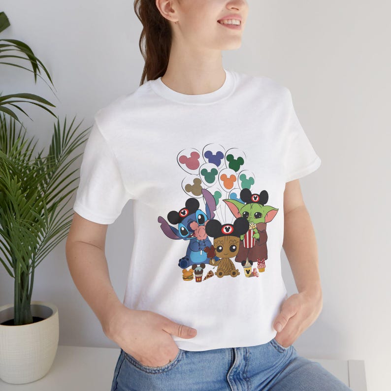 Puede incluir: Camiseta blanca con un estampado gr&aacute;fico de Stitch, Baby Yoda y Groot sosteniendo globos de Mickey Mouse. Los personajes llevan orejas de Mickey Mouse.