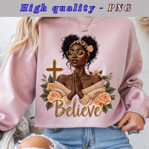Puede incluir: Una sudadera rosa claro con un gráfico de una mujer negra con los ojos cerrados, con gafas y una diadema con estampado de leopardo, con la palabra "Believe" en escritura dorada. El diseño incluye una cruz y rosas color melocotón.