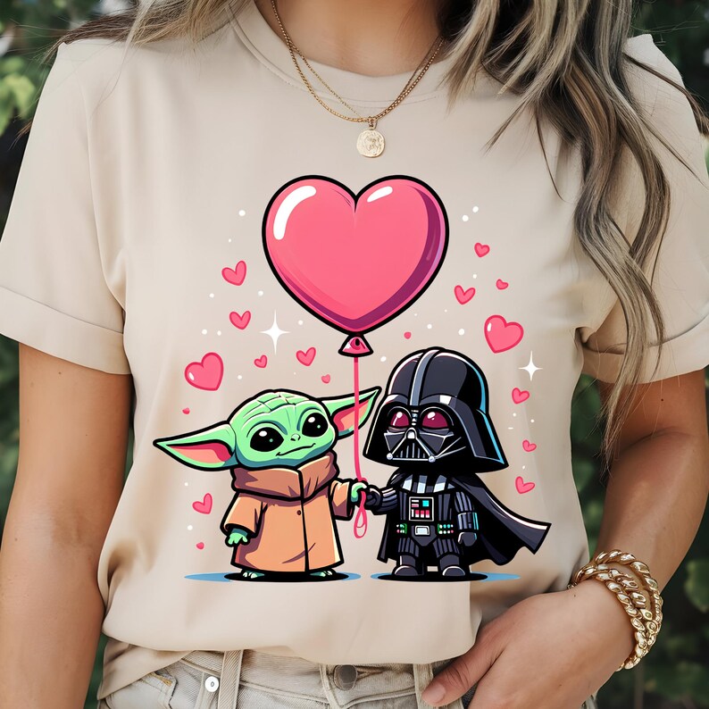 May include: Beige t-shirt with a cartoon illustration of Baby Yoda and Darth Vader holding hands and a pink heart-shaped balloon. Pink hearts and white dots are scattered around the characters. The shirt is a light tan colour.