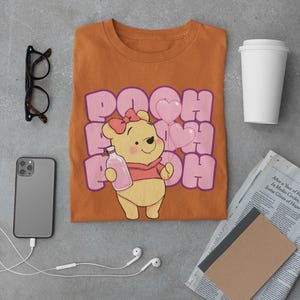 May include: A brown t-shirt with a cartoon image of Winnie the Pooh holding a bottle and pink heart balloons. The text "POOH" is printed in pink and purple letters.