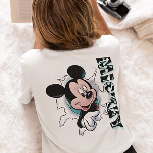 以下が含まれることがあります： 白いTシャツに、穴から顔を出したミッキーマウスのグラフィックと、横に「MICKEY」と縦書きで書かれた文字がプリントされています。