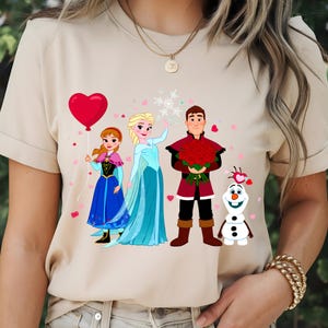 Pu&ograve; includere: T-shirt beige con un'illustrazione di personaggi di un popolare film d'animazione. Il design include un palloncino a forma di cuore, fiocchi di neve e coriandoli a forma di cuore. I personaggi indossano abiti colorati.