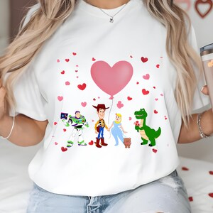 May include: White t-shirt featuring cartoon characters Buzz Lightyear, Woody, Cinderella, and Rex, with a pink heart balloon and scattered hearts. The design is a Valentine's Day theme.