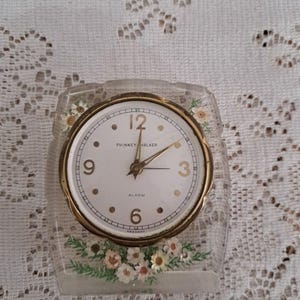 May include: A vintage Phinney Walker alarm clock with a clear, rectangular case. The clock face is white with gold numerals and hands. The case is decorated with painted flowers. The clock reads "ALARM" and "GERMANY".