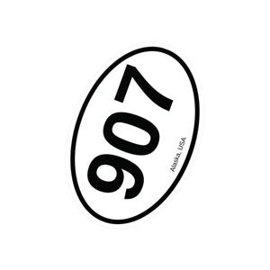 May include: Oval sticker with a black border, featuring the number "907" in bold black font. The text "Alaska, USA" is printed in a smaller font along the bottom edge of the oval. The sticker is white.