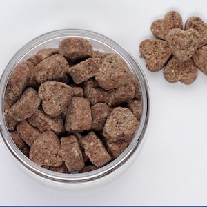 K&ouml;nnte beinhalten: Eine Glassch&uuml;ssel gef&uuml;llt mit herzf&ouml;rmigen braunen Hunde-Leckerlis. Die Leckerlis sind in einem Haufen angeordnet und leicht uneben. Neben der Sch&uuml;ssel befinden sich auch ein paar herzf&ouml;rmige Leckerlis auf einer wei&szlig;en Oberfl&auml;che.