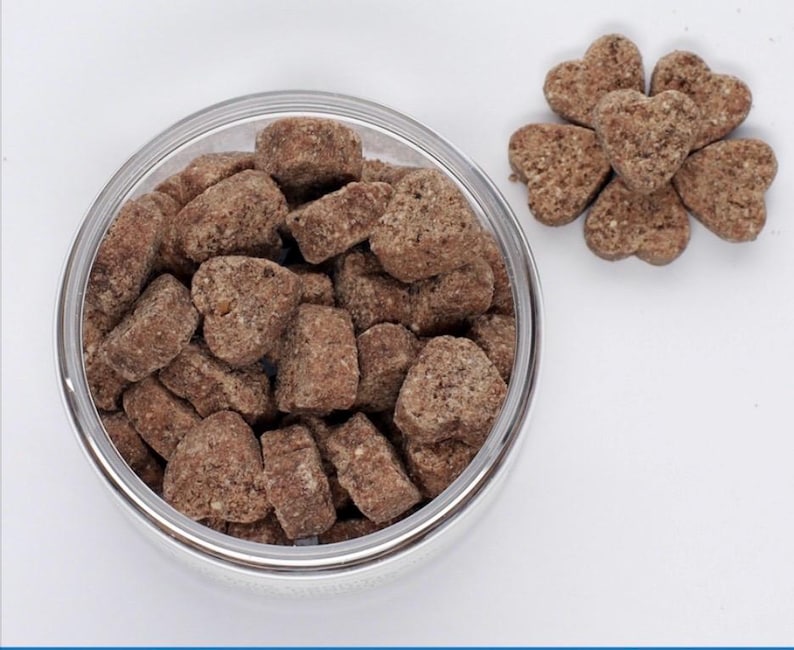 K&ouml;nnte beinhalten: Eine Glassch&uuml;ssel gef&uuml;llt mit herzf&ouml;rmigen braunen Hunde-Leckerlis. Die Leckerlis sind in einem Haufen angeordnet und leicht uneben. Neben der Sch&uuml;ssel befinden sich auch ein paar herzf&ouml;rmige Leckerlis auf einer wei&szlig;en Oberfl&auml;che.