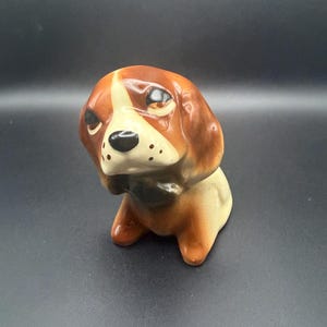 Puede incluir: Figura de perro de cerámica en tonos marrón, beige y negro. El perro tiene una expresión inquisitiva, con una raya blanca en la cara y una nariz negra. La figura es un objeto decorativo.