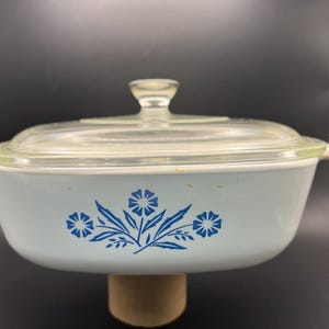 Puede incluir: Una fuente para horno blanca con un diseño floral azul y una tapa de vidrio transparente. La fuente tiene forma redonda y un borde ligeramente elevado.