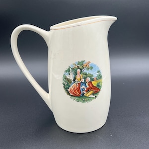 May include: Cream-colored ceramic pitcher with a curved handle and a spout. A circular image on the front depicts a couple in period clothing in a garden setting. The rim has a gold-colored trim.