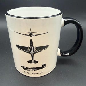 May include: White ceramic mug with a black handle and rim. The mug features black illustrations of three different airplanes, including a P-40E Warhawk. The mug is a standard size for coffee or tea.