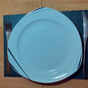 Könnte beinhalten: Ein weißer Teller mit schwarzen Linien am Rand liegt auf einem schwarzen Schieferuntersetzer mit einer silbernen Gabel und einem Messer.