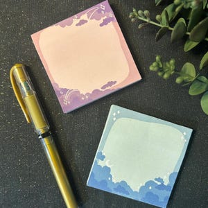 May include: Two square sticky note pads with a pastel pink and blue sky design. The pink pad has white stars and clouds with a rainbow. The blue pad has white stars, clouds, and a crescent moon. Both pads have a blank space for writing.