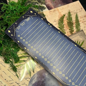 May include: A dark blue To Do List notepad with gold star and diamond accents. The notepad has lined pages and the words "To Do List" at the top. The notepad is surrounded by vintage paper, moss, and moon-shaped decorations.