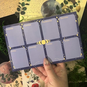 May include: A celestial-themed weekly planner pad with a dark blue background and light blue boxes for each day of the week. Gold stars and constellation designs decorate the edges. The days of the week are printed in gold.