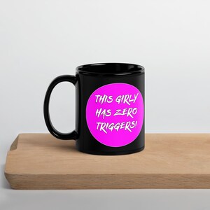 Può includere: Una tazza in ceramica nera con manico bianco. La tazza presenta un cerchio rosa acceso con la scritta "THIS GIRLY HAS ZERO TRIGGERS!" in bianco, in grassetto e maiuscolo. La tazza è appoggiata su una superficie di legno.