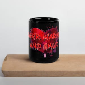 Può includere: Tazza in ceramica nera con un design a forma di cuore rosso e le parole "ANGER WARN AND SMUT" in lettere rosse. Il cuore ha un effetto acquerello con accenti viola.