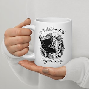 May include: White ceramic mug featuring a black and white illustration of a skeleton reading a book. The mug has the text "My Books Come With Trigger Warnings" in a gothic font. The design includes floral elements.