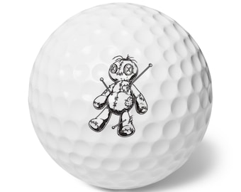 May include: White golf ball with a black and white illustration of a voodoo doll. The doll has stitched features, pins, and a patch. The golf ball has a dimpled surface.