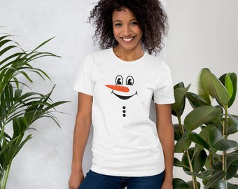 May include: White t-shirt featuring a snowman face design. The design includes black eyes, a carrot nose, a smiling mouth, and three black buttons. The shirt is worn with blue jeans.
