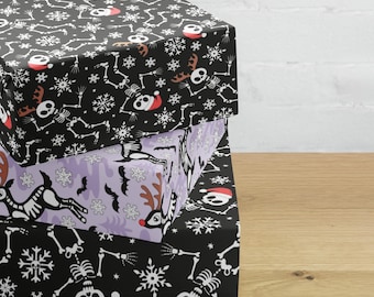 May include: Three stacked square gift boxes with festive Halloween and Christmas patterns. The top box is black with white skeleton and snowflake designs, and red Santa hats. The middle box is lavender with reindeer and bat designs. The bottom box is black with skeleton and snowflake designs.