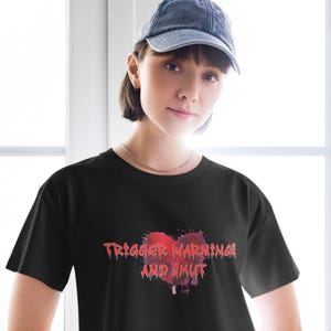 Puede incluir: Camiseta negra con un gráfico de corazón rojo y el texto "TRIGGER WARNINGS AND SMUT" en una fuente distorsionada. La camiseta es de cuello redondo y la lleva una persona con una gorra de béisbol vaquera.