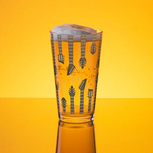 Può includere: Un bicchiere da pinta trasparente pieno di birra e schiuma, con un motivo di paletta di chitarra nera. Il bicchiere è posto su uno sfondo giallo brillante, creando un'immagine vibrante.
