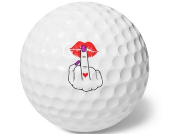 May include: White golf ball featuring a graphic of red lips, a middle finger, and a heart. The middle finger has a purple fingernail. The lips and heart are red, and the fingernail is purple. The golf ball has a dimpled surface.