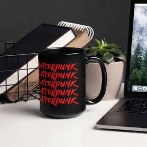May include: A black coffee mug with the word "Latterpunk" in red, stacked multiple times. The mug is on a white surface, with a laptop, a succulent plant, and a wire basket with notebooks in the background.