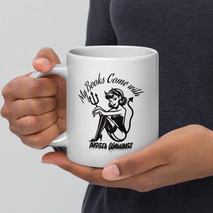 May include: White ceramic mug featuring a black illustration of a devil woman with the text "My Books Come with Trigger Warnings." The devil woman is seated, holding a trident. The mug has a curved handle.