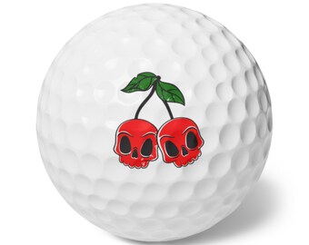 May include: White golf ball with a design of two red skull-shaped cherries and green leaves. The ball has a dimpled surface.