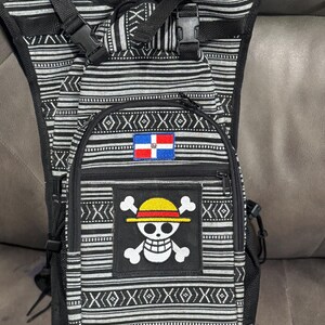 May include: Black and white patterned backpack with adjustable straps and a front zipper pocket. Features a patch with a skull and crossbones wearing a yellow hat, and a Dominican Republic flag patch.