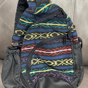May include: A small, patterned backpack with a black base and mesh side pockets. The fabric features a colorful geometric design with blue, red, yellow, and purple accents. The bag has multiple zippered compartments and adjustable straps.