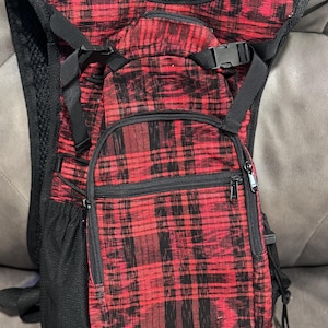 May include: Red and black plaid backpack with multiple zippered compartments and adjustable straps. The backpack has a black mesh back panel and a chest buckle. The design features a classic plaid pattern, suitable for outdoor activities or everyday use.