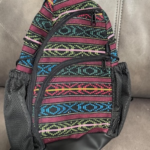 May include: A colorful sling backpack with a geometric pattern in shades of red, black, blue, yellow, and pink. The bag has two zippered compartments, a black mesh side pocket, and a black bottom. The bag is designed to be worn over one shoulder.