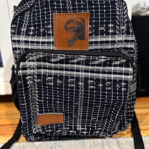May include: Black and white patterned backpack with a brown leather patch featuring a black and white illustration of Venom.