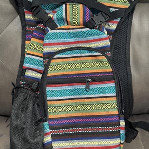 May include: A colorful backpack with a vibrant, patterned design. The backpack features a variety of horizontal stripes in red, orange, blue, and green, with geometric accents. It has a small front pocket, a mesh side pocket, and adjustable straps.