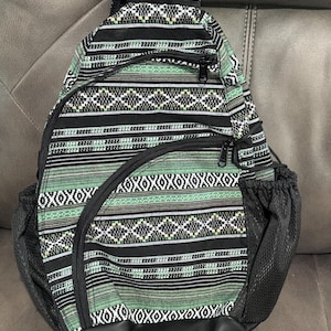 May include: A black and green patterned sling backpack with a black mesh side pocket and a black bottom. The backpack has a geometric design with white accents and black zippers. The bag has a black strap and is made of a durable fabric.