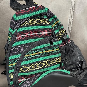 May include: A small, black and patterned sling backpack with a black base and mesh side pocket. The fabric features a repeating pattern of green, black, red, yellow, and purple horizontal stripes and geometric shapes. The bag has a black zipper and strap.