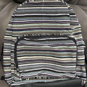 May include: A backpack with a striped pattern in black, white, purple, blue, and tan. The backpack has a front pocket with a zipper and a black bottom. The straps are black.