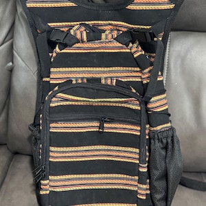 May include: Black backpack with horizontal stripes in orange, yellow, and red. The bag has a front zippered pocket, two side mesh pockets, and adjustable straps. The backpack is designed to be worn across the chest.