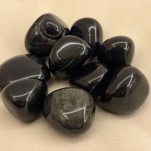 Könnte beinhalten: Eine Ansammlung polierter schwarzer Obsidiansteine. Die Steine sind glatt, abgerundet und glänzend, wodurch sie das Licht reflektieren. Die Formen variieren leicht, einige zeigen subtile innere Muster.