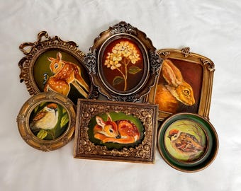 May include: A collection of framed vintage paintings featuring wildlife. The artwork includes a fawn, a rabbit, and birds, all set against a dark background. The frames are ornate, with gold and bronze tones, adding to the antique aesthetic.