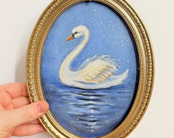 May include: An oval-shaped painting of a white swan swimming on blue water, set within an ornate gold-colored frame. The swan is depicted in profile against a blue background with white speckles, and its reflection is visible on the water surface.
