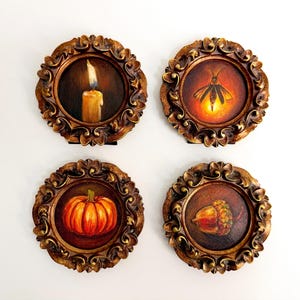 Peut inclure: Quatre plaques murales circulaires avec des cadres dorés antiques ornés. Chaque plaque présente une image peinte distincte : une bougie allumée, une luciole brillante, une citrouille orange et un gland brun, sur des fonds sombres et chauds.