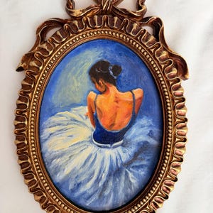 以下が含まれることがあります： 金色のフレームに入ったバレリーナの楕円形の絵画。 バレリーナは後ろ姿で、青いレオタードと白いチュチュを着ています。 フレームの上部には装飾的なリボンがあり、詳細なボーダーがあります。