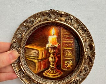 May include: A small, round, framed painting depicts a candle in a gold candlestick, books, and a warm, inviting glow. The painting is in a gold ornate frame, creating a vintage aesthetic. The artwork evokes a cozy, scholarly theme.