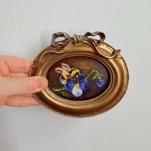 May include: Oval-shaped, antique gold-colored frame with a miniature painting of a bumblebee on a blue and white flower. The frame has a decorative bow at the top and a beaded edge. The painting is set against a dark brown background.