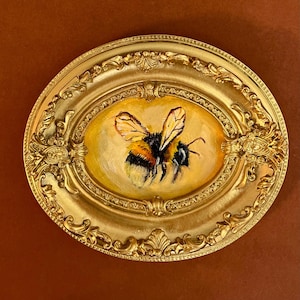 May include: A gold-coloured, oval frame with intricate detailing encases a painting of a bee. The bee is painted with black and yellow stripes, and its wings are a translucent yellow. The painting's background is a warm yellow.